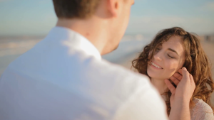 young-man-touch-face-of-his-woman-young-caucasian-couple-on-the-seashore-wind-play-in-her-hair-woman-look-at-her-partner-and-smile