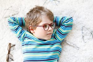 Close-up portrait of little blond kid boy with different eyeglasses on white background. Happy smiling child in casual clothes. Childhood, vision, eyewear, optician store. Boy choosing new glasses.
